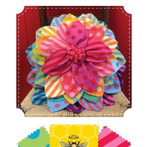 May include: A colorful, flower-shaped pillow with the text "Petal Pop Zinnia Pillow." The pillow features various patterns, including polka dots and stripes, in shades of pink, orange, yellow, and blue. The pillow is sitting on a woven chair.