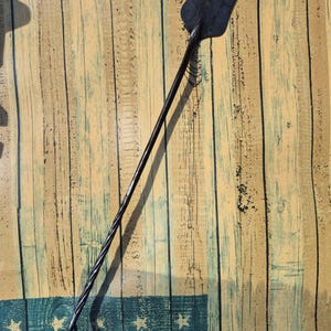 May include: A long, black, hand-forged metal fire poker featuring a flat, spade-shaped head at one end and a twisted, textured handle that terminates in a curved hook. The tool has a dark, matte finish and a slender, elongated design.