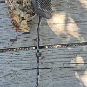 May include: A black grill brush with a square head and metal bristles. The handle is twisted and has a hook at the end. The brush is designed for cleaning grills and barbecues.