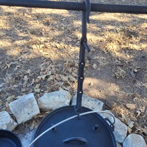 May include: Black cast iron Dutch oven with a lid hanging from a metal hook. The oven is sitting on a bed of rocks. The text "Camp Chef" is visible on the front of the oven.