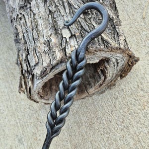 May include: Hand-forged metal hook with a twisted handle and a curved hook end. The dark gray metal contrasts with the textured, light brown wood in the background. This rustic piece is likely for hanging items.