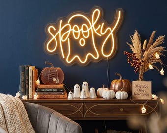 May include: A yellow neon sign shaped like a ghost with the word spooky written in cursive script. The sign is mounted on a dark blue wall above a wooden console table decorated with books, small ghost figurines, and pumpkins.
