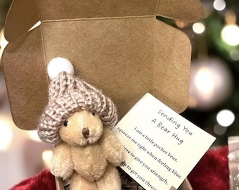 May include: A small brown teddy bear wearing a knitted hat is in a brown box with a white card that says "Sending You A Bear Hug. I am a little pocket bear, squeeze me tight when feeling blue. Here to give you strength, help to get you through."