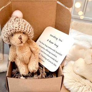 May include: A small, tan teddy bear wearing a knitted hat with a pom-pom, presented in a cardboard box. A card reads "Sending You A Bear Hug" with a message of comfort. The box contains shredded paper for cushioning.