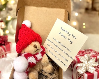 May include: A small, red and white knitted bear wearing a red scarf is sitting in a brown cardboard box with shredded paper. The bear is holding two white pom-poms. A card with the text "Sending You A Bear Hug" is visible in the box.  I am a little pocket bear, squeeze me tight when feeling blue I am here to give you strength, that will help to get you through.