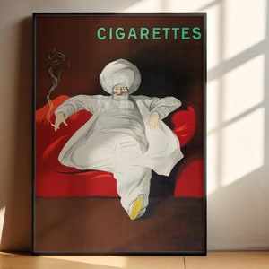 May include: Vintage poster print featuring a seated figure in white robes and a turban, holding a cigarette. The word "CIGARETTES" is in green at the top. The figure is seated on a red cushion against a dark brown background, framed in black.