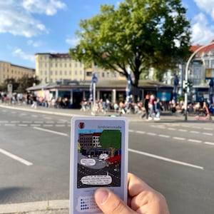 May include: A card game with a cartoon illustration of a street scene with the text "Sch&ouml;nleinstra&szlig;e" and "COOL YOLL PLATI" and "ANANAS, MELONE, PIRSICA EGAL WAS, FOR SA HEUTE ALLES 1&euro;". The card has a scale of 100, 200, 1000, 2000.