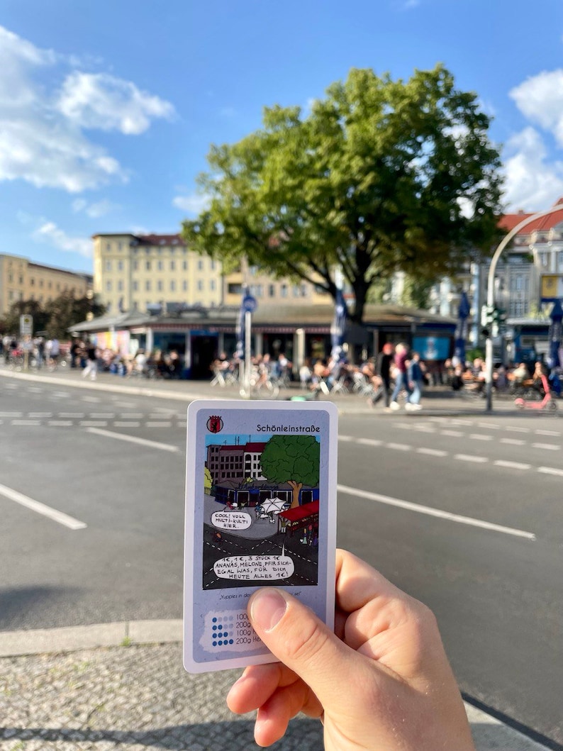 May include: A card game with a cartoon illustration of a street scene with the text "Sch&ouml;nleinstra&szlig;e" and "COOL YOLL PLATI" and "ANANAS, MELONE, PIRSICA EGAL WAS, FOR SA HEUTE ALLES 1&euro;". The card has a scale of 100, 200, 1000, 2000.