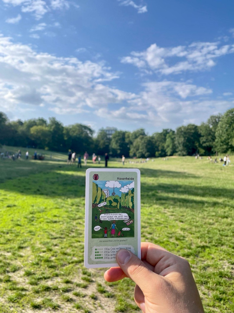May include: A green and white card with a cartoon illustration of a park with a tower in the background. The card has the title "Hasenheide" and the text "MAMAAN DEJ! Dann gehen wir jetzt mal in den SCHLOSS STEIGERN!" and "und second floor und die gehen!" The card also has a scale with the text "100g Gras mit 100E", "200g Gras mit 200E", "500g Gras mit 500E", and "1000g Gras mit 1000E".