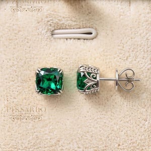 May include: A pair of stud earrings featuring square-cut, deep green gemstones set in silver-toned metal. The side of the setting displays an intricate, openwork filigree pattern. The earrings are presented on a cream-colored velvet display pad.