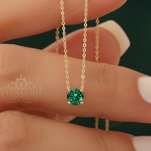 Puede incluir: Un delicado collar de oro con un vibrante colgante de piedra preciosa verde. La esmeralda, de talla redonda, está engastada en un diseño sencillo y elegante. El collar se muestra sobre un fondo suave y borroso, resaltando el brillo y el color de la joya.
