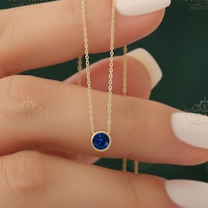 Puede incluir: Un delicado collar de cadena dorada con un colgante de una sola piedra preciosa azul redonda engastada en bisel. El collar se sostiene verticalmente, mostrando los eslabones dorados pulidos y la piedra circular de color azul intenso.