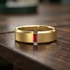 May include: A gold ring with a brushed texture, featuring a rectangular, red gemstone. The ring is set on a dark wooden surface. The ring is a piece of jewellery.