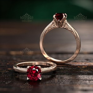 May include: Two gold rings with a large, round, deep red gemstone. The rings have intricate floral designs on the band and setting. The rings are displayed on a dark wooden surface.