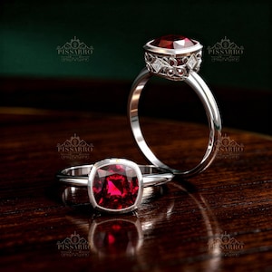 May include: Two silver rings with large, square, deep red gemstones. One ring is in focus, showcasing the gemstone's facets and a detailed setting. The other ring is slightly out of focus, with a similar design. The rings are displayed on a dark wooden surface.