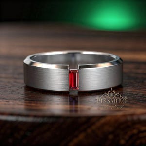 May include: A silver ring with a brushed finish and a rectangular, vibrant red gemstone. The ring has a modern design with a recessed channel holding the gemstone. The ring is displayed on a wooden surface.