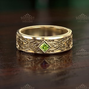 May include: A gold-colored ring with an ornate, engraved design. The ring features a square, light green gemstone at the center. The ring's band is detailed with a repeating floral pattern. The ring is displayed on a dark wooden surface.