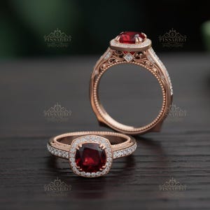May include: Two rose gold rings with a square, deep red gemstone center, surrounded by smaller white gemstones. The bands feature intricate filigree detailing. The rings are displayed on a dark wooden surface.