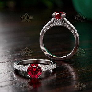 May include: Two silver rings with red gemstones. One ring features a large, round ruby center stone, while the other has a smaller ruby. Both rings have a band adorned with small, clear gemstones. The rings are displayed on a dark wooden surface.