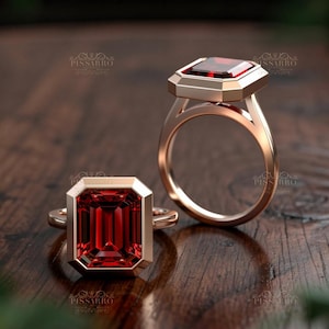 May include: Two rose gold rings with large, rectangular, deep red gemstones. The gemstones are set in a bezel setting. The rings are displayed on a wooden surface. The rings are elegant and eye-catching.