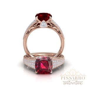 Könnte beinhalten: Zwei roségoldene Ringe mit großen, roten Edelsteinen. Ein Ring hat einen kissenförmigen Stein, während der andere einen runden Stein hat. Beide Ringe sind mit kleinen, klaren Akzentsteinen und aufwendigen Details verziert.