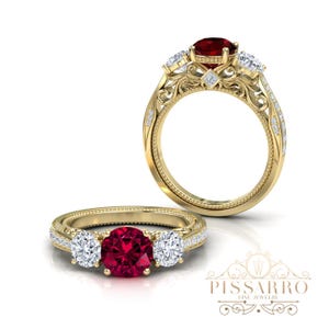 May include: A gold engagement ring with a large, round, red gemstone center and two smaller white gemstones on either side. The ring features intricate filigree detailing and small accent stones. The brand name "Pissarro Fine Jewelry" is visible.