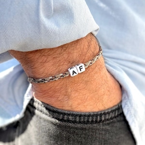Puede incluir: Una pulsera plateada con un diseño tejido y cuentas blancas con las letras "AF". La pulsera se lleva en la muñeca, combinada con una camisa azul claro y vaqueros grises oscuros. La pulsera es una joya.