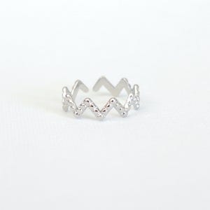 Könnte beinhalten: Ein silberfarbener Ring mit Zickzack-Muster. Der Ring hat eine strukturierte Oberfläche und eine Öffnung oben zur Größenanpassung. Er wird vor einem weißen Hintergrund präsentiert, was sein schlichtes, aber stilvolles Design hervorhebt.