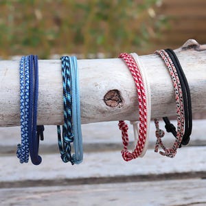 Peut inclure: Une collection de six bracelets en corde de différentes couleurs, dont le bleu, le noir, le rouge et le blanc. Les bracelets sont présentés sur une branche en bois clair, mettant en valeur leurs motifs tissés. L'arrière-plan est flou.