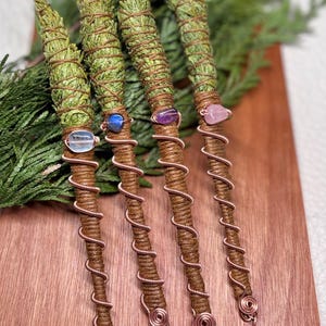 May include: Four sage smudge sticks wrapped in brown twine and copper wire. Each stick features a different gemstone: clear quartz, blue, purple, and pink. The sticks are displayed on a wooden surface with green foliage in the background.