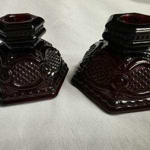 Peut inclure: Deux encriers anciens en verre de couleur rubis. Chaque encrier a un dessus carré et étagé et un corps décoratif à motifs de losanges. Les encriers sont placés sur une surface blanche. Le verre a une finition brillante.