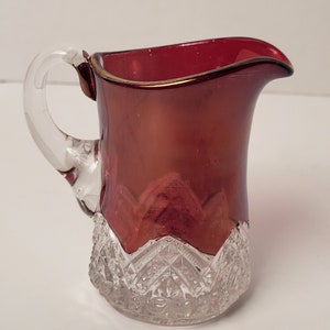 May include: A ruby red glass creamer with a clear glass handle. The creamer has a gold rim and a diamond-cut pattern at the base. The creamer is a vintage style and is likely used for serving cream or other liquids.