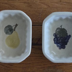 Könnte beinhalten: Zwei cremeweiße Keramikschalen mit gewellten Rändern. Eine Schale zeigt eine gelbe Birne mit einem grünen Blatt, die andere eine Traube aus lila Trauben. Die Schalen sind rechteckig mit abgerundeten Ecken.