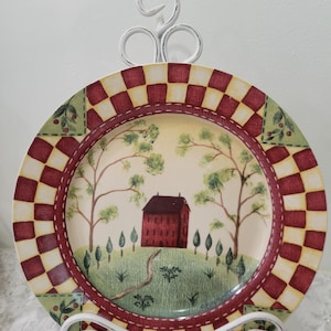 May include: A decorative plate with a rural scene featuring a red building, trees, and a path. The plate has a red and yellow checkered border and is displayed on a white metal stand. The plate's design evokes a rustic, country theme.