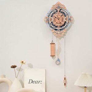 May include: A decorative wooden wall clock with a light blue and tan face, Roman numerals, and ornate detailing. The clock has a pendulum and decorative accents. The word "Dear:" is printed on a white card.