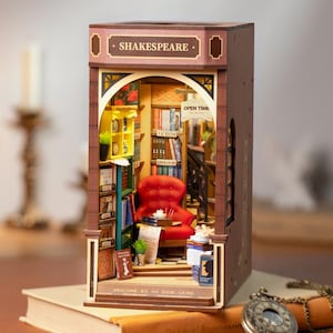 Op de afbeelding: Een gedetailleerd miniatuur houten model van een boekwinkel, met de naam "Shakespeare" boven de ingang. Binnenin staat een rode fauteuil tussen de boekenplanken. De tekst "Welcome to my book store" is zichtbaar.