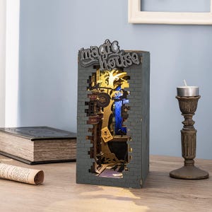 Op de afbeelding: Een grijze, houten "magic house" boekenhoek met een bakstenen muurontwerp. Het interieur toont een gedetailleerde, verlichte sc&egrave;ne met een boom, borden en een blauw raam. Een kandelaar en een boek zijn op de achtergrond.