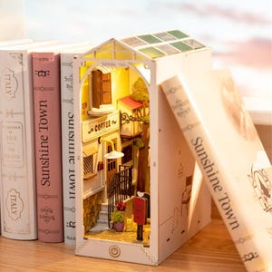 Op de afbeelding: Een miniatuur diorama van een Europese straatsc&egrave;ne met een caf&eacute;, trappen en een rode brievenbus. Het verlichte diorama staat tussen boeken met de titel "Sunshine Town". De sc&egrave;ne staat op een houten oppervlak.