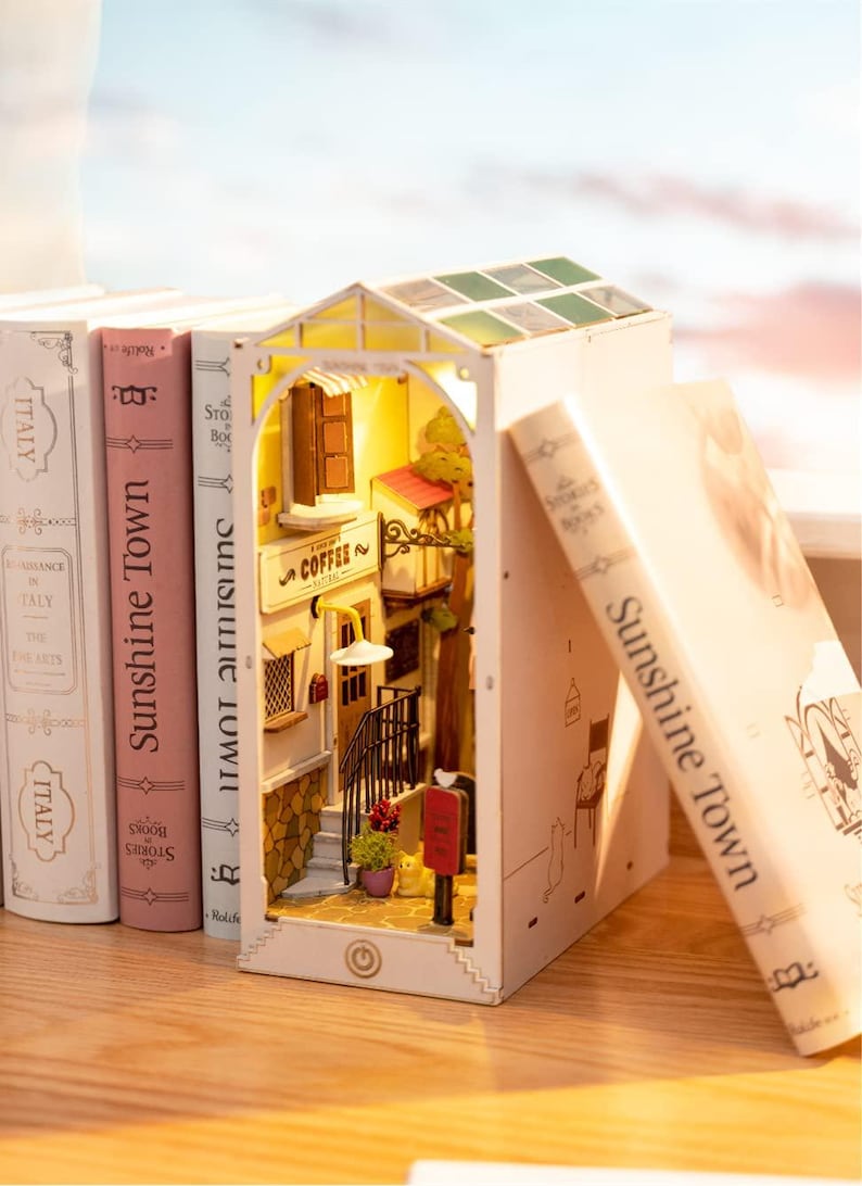 Op de afbeelding: Een miniatuur diorama van een Europese straatsc&egrave;ne met een caf&eacute;, trappen en een rode brievenbus. Het verlichte diorama staat tussen boeken met de titel "Sunshine Town". De sc&egrave;ne staat op een houten oppervlak.