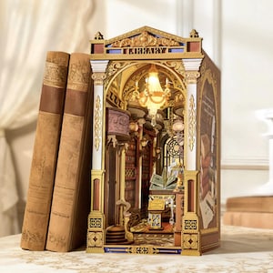 May include: A detailed miniature library diorama with ornate gold and brown architectural details. The word "LIBRARY" is displayed above the entrance. Three antique books lean against the diorama, creating a layered effect. The interior features bookshelves, a chandelier, and a reading table.