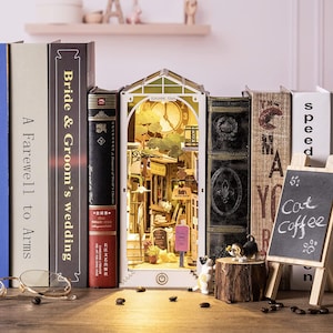 Op de afbeelding: Een gedetailleerd miniatuur stadsmodel, verlicht van binnen, staat tussen boeken. Het model toont een klokkentoren en winkelpuien. Een klein schoolbordje toont "Cat Coffee". Decoratieve beeldjes en koffiebonen zijn in de buurt geplaatst.