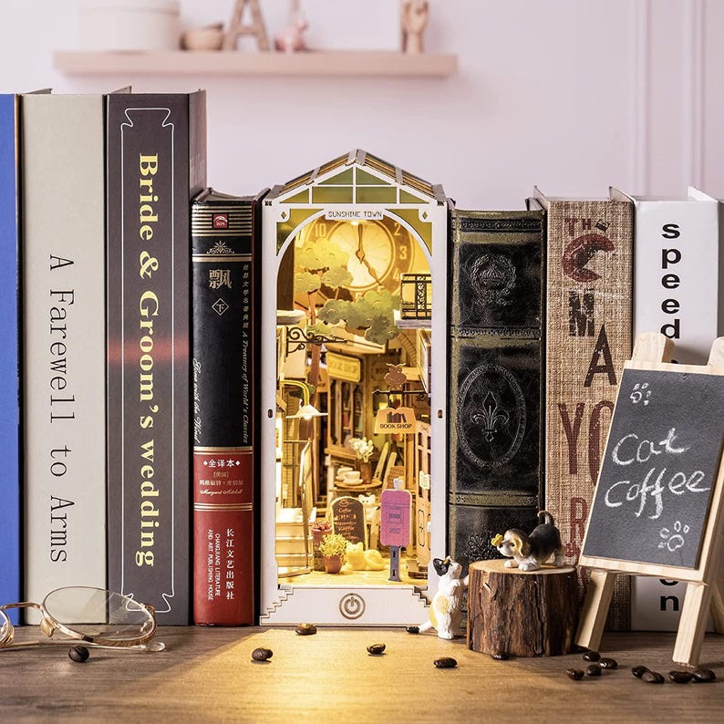 Op de afbeelding: Een gedetailleerd miniatuur stadsmodel, verlicht van binnen, staat tussen boeken. Het model toont een klokkentoren en winkelpuien. Een klein schoolbordje toont "Cat Coffee". Decoratieve beeldjes en koffiebonen zijn in de buurt geplaatst.