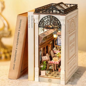 Op de afbeelding: Een miniatuur diorama van een caf&eacute;sc&egrave;ne, geconstrueerd in een boek. De rug van het boek leest "Dr. Bob Arnot's WEIGHT CONTROL PROGRAM". Het diorama heeft een gedetailleerd interieur met tafels, stoelen en decoratieve elementen. De boekomslag leest "ARCADE DATING".