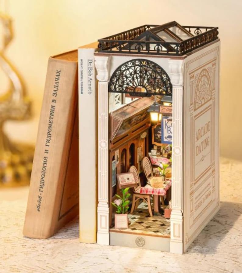 Op de afbeelding: Een miniatuur diorama van een caf&eacute;sc&egrave;ne, geconstrueerd in een boek. De rug van het boek leest "Dr. Bob Arnot's WEIGHT CONTROL PROGRAM". Het diorama heeft een gedetailleerd interieur met tafels, stoelen en decoratieve elementen. De boekomslag leest "ARCADE DATING".