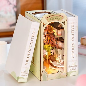 Op de afbeelding: Een houten boekenhoek met een miniatuur Japans dorpsgezicht, verlicht door warm licht. De boekenhoek wordt geflankeerd door twee witte boekomslagen met de tekst "Falling Sakura" en "Stories in Books". De sc&egrave;ne omvat miniatuurgebouwen, bomen en een trap.