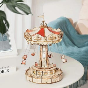 May include: A wooden carousel model with a red and white striped canopy and small swings. The carousel has intricate details and is illuminated with small lights. The base and supporting structure are made of light-colored wood, creating a charming decorative piece.