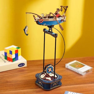 May include: A wooden model of a fishing boat with a blue and white hull, featuring miniature figures and a fishing rod with a fish. The boat is suspended above a detailed base with a world map design. A Rubik's cube and a cassette tape are also visible.