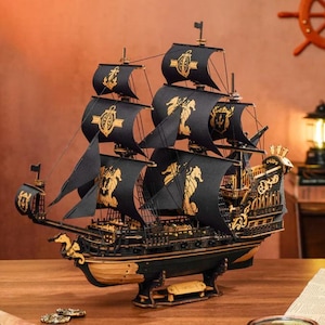 May include: A detailed wooden model of a black pirate ship with gold accents. The ship features black sails adorned with gold seahorses and crests. The model is displayed on a wooden surface, showcasing intricate craftsmanship and design.