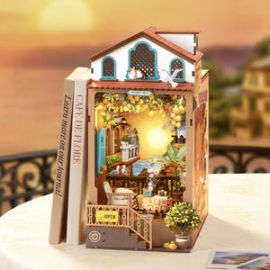Könnte beinhalten: Ein Miniatur-Bücherregal-Einsatz aus Holz mit einer mediterranen Balkonszene, Sonnenuntergang, Zitronenbäumen und einer kleinen Katze. Die Struktur umfasst eine weiße Hausfassade, ein gelbes 'OPEN'-Schild und ein Power-Button-Symbol am Sockel.