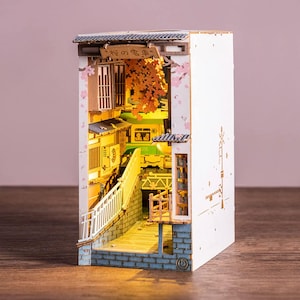 Op de afbeelding: Een miniatuur houten diorama van een Japanse straatsc&egrave;ne, verlicht met warme verlichting. De sc&egrave;ne omvat een brug, gebouwen en kersenbloesem accenten. Het diorama heeft de Japanse tekst "桜の電車" op een bord.